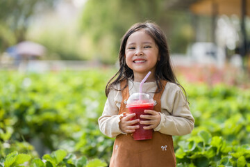 Naklejka premium happy preschool child girl holding a cup of fresh strawberry smoothie in farm
