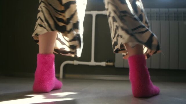 Joyful woman in fluffy pink socks and tiger print pants dancing barefoot on floor at home, with sunlight casting shadows across room, creating playful and carefree mood. Woman legs dancing at home