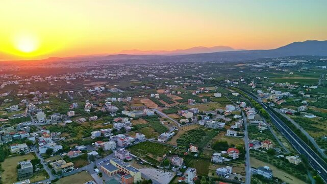Drone shot over Heraklion, Crete at sunset, showing roads, fields and low-rise neighborhoods. Warm evening light spreads across the urban fringe and surrounding landscape.