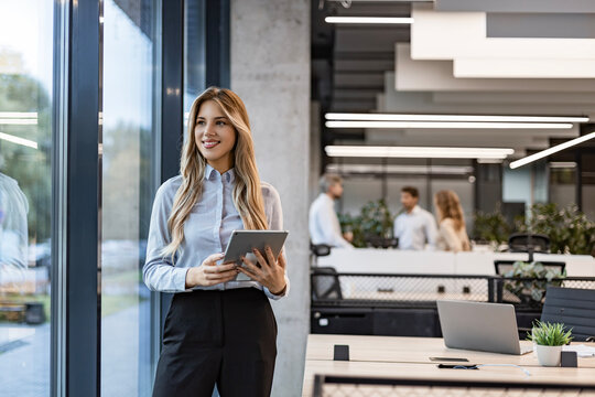 Young happy businesswoman using digital tablet while standing by the window in the office and looking at camera