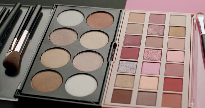 Assortment of cosmetic products including eyeshadow and highlighter palettes with various shades, accompanied by a set of professional makeup brushes on a pink and black background