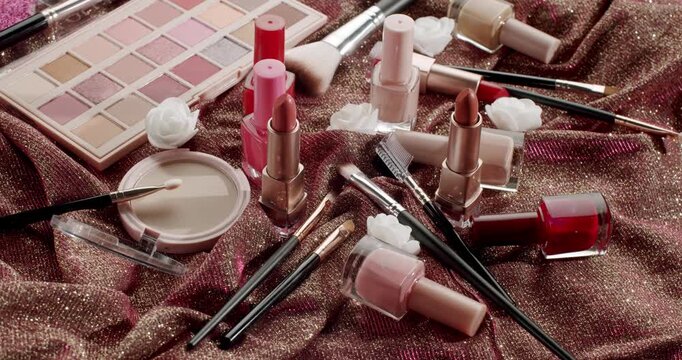 Assortment of decorative cosmetic products including eyeshadow palette, lipsticks, nail polishes, powder, and brushes lying on a shiny pink fabric, perfect for a glamorous beauty routine