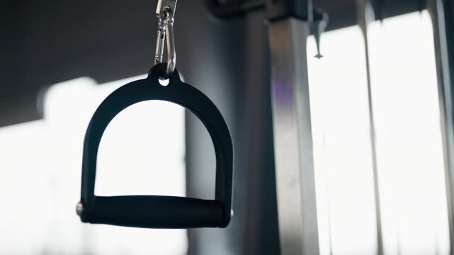 Detailed close-up shot of a single handle of a crossover or lat pulldown cable machine swinging in a modern gym with bright windows in the background, showing equipment ready for a workout