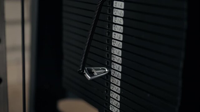 Close-up of a hand inserting a pin to select the weight on a modern gym machine weight stack, preparing for a strength training workout in a dark and moody fitness center in slow motion