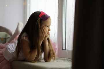 Naklejka premium Young Girl in Princess Costume Daydreaming by Window, Wearing Purple Mask, Children's Imagination, Indoors