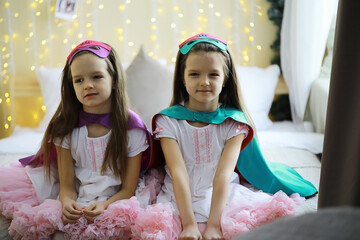 Naklejka premium Twin Girls in Princess Costumes with Capes and Masks Sitting on Bed with Fairy Lights