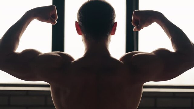 Silhouette of a muscular man with a bare torso posing in front of a window, showcasing his strength and perfect physique as he slowly flexes his biceps in a classic double biceps pose