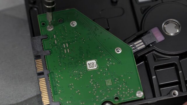 A person is taking apart a hard drive using a screwdriver. They carefully remove screws and handle the parts. Tools and a work surface are visible in the background.