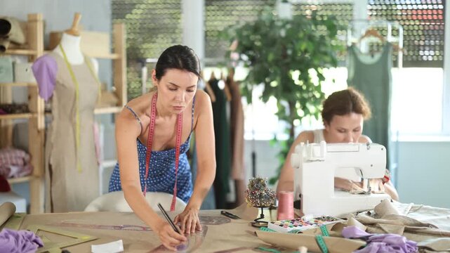 Woman tailor draw dress pattern with curve ruler on paper, female master seamstress sewing cloth with sewing machine. Tailor workplace with cut textile, sewing machine, thread, pins, needles, tape