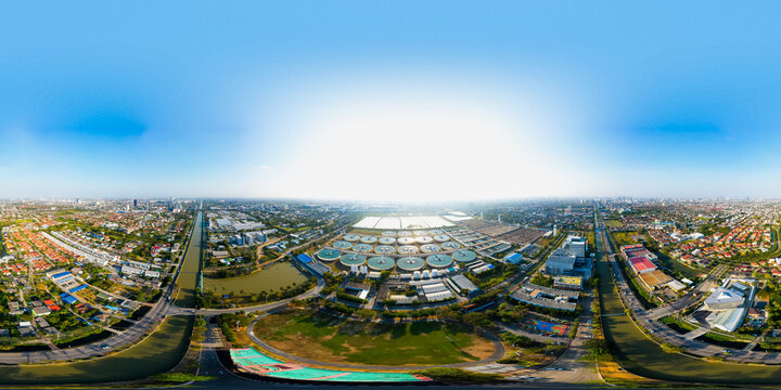 Ultra panoramic 360 aerial view of smart city wastewater treatment plant with circular clarifier tanks surrounded by urban landscape, showcasing sustainable water management under bright daylight.