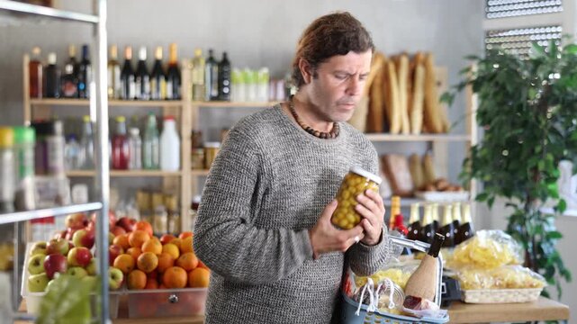 Man customer with shopping cart or basket buying food in supermarket grocery store, reading ingredients on jar of conserve olives