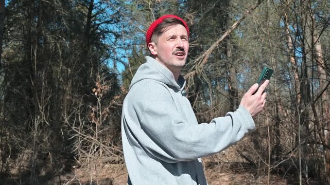 A man looks at his smartphone while standing outdoors in the forest. He's looking for a connection or checking his geolocation while lost during a nature walk