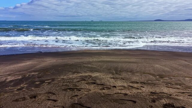 Waves move onto a sandy beach with a cloudy sky above. Distant ships are seen on the horizon, creating a sense of openness and space. The scene captures a coastal atmosphere.