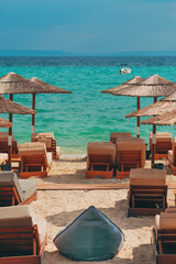 Fototapeta premium Sandy beach with lounge chairs and straw umbrellas facing turquoise sea. Relaxing coastal scene with calm water and distant boat under a clear sky. Mediterranean summer setting.