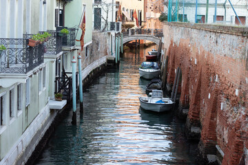 Narrow canal in Venice with boats moored along the brick walls and a small arch bridge connecting the buildings on either side of the waterway