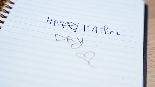 lined spiral notebook showing a handwritten Happy Father Day message with a heart, placed on a wooden desk indoors with soft focus and natural detail.