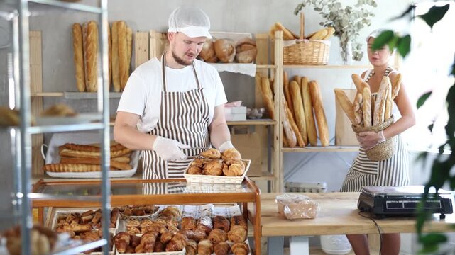 Smiling young adult female worker puts croissants in the case as her colleague stacks baguettes
