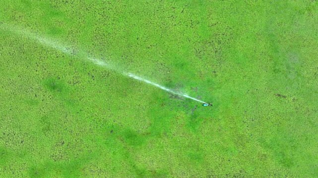 Drone shot of a lawn being watered evenly by a sprinkler, highlighting modern landscape management, efficient resource use, and consistent care for healthy grass development.
