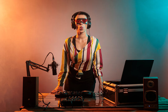 Female artist sticking tongue out and being funky, acting silly and fooling around over studio background. Confident cool DJ using turntables and audio equipment, mixing music.