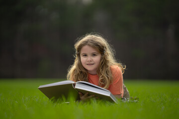 Naklejka premium Kid read a book in the grass. Kid read a book story outdoors. Kid read book in the meadow on a sunny day. Reading in nature. Schoolboy with book. Kid outdoor learning. Kids read story.