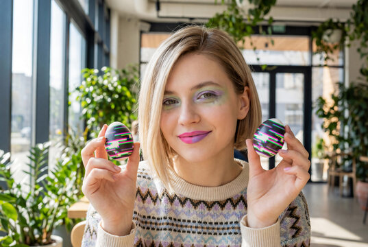 Young woman with asymmetrical lob and eccentric make-up holding easter egg with digital glitch pattern in bright cafe interior for avant garde make-up editorial concept