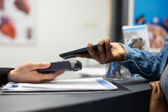 Closeup of contactless payment transaction as african american patient taps phone at hospital front desk. Clinic receptionist holds pos as client uses nfc technology for secure, cashless checkout.
