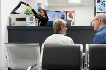 Naklejka premium Asian receptionist using copier and scanning machine at front desk of modern healthcare facility. Female hospital secretary handles printing of medical forms as elderly doctor consults with patient.