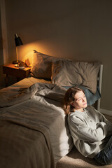 Fototapeta premium Vertical of Caucasian young adult woman sitting on floor beside bed gazing thoughtfully into distance, sunlight illuminating face, expressing emotion of contemplation or introspection
