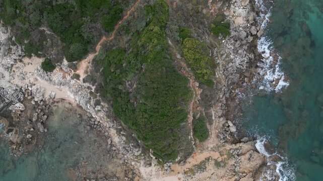 Aerial view of a rugged coastline with green vegetation, rocky shores, sandy patches, and turquoise waters with gentle waves.