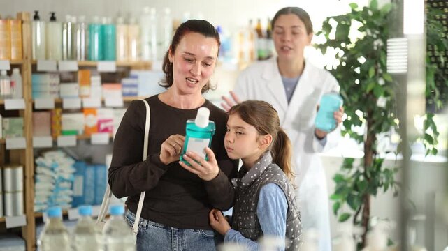 Woman with curious preteen daughter inspecting bottle of oral rinse at local pharmacy while smiling female pharmacist in background offering advice and other product to compare. High quality 4k