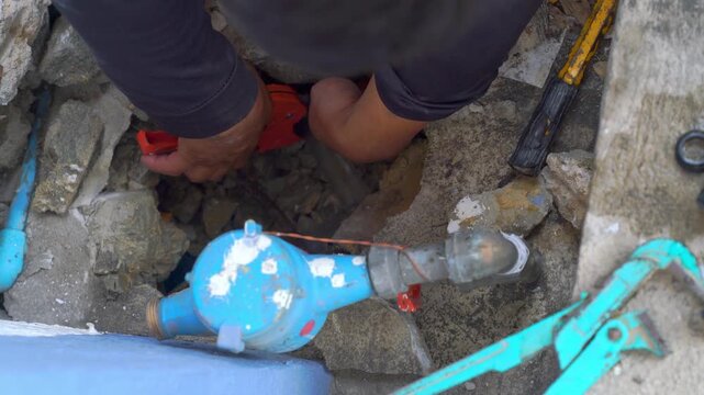 Plumber hands repairing underground pipe leak through concrete opening. High angle view showing blue water meter, pipe wrench, and hammer tools. Concept of home maintenance and utility repair work.