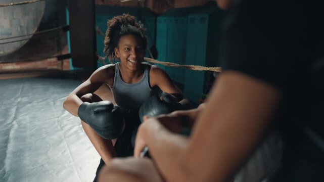 Happy young athletic lady talking cheerfully with coach in gym, sit on box ring. Professional sport and self-defense class for women, hobby for good body shape and endurance development, good mood