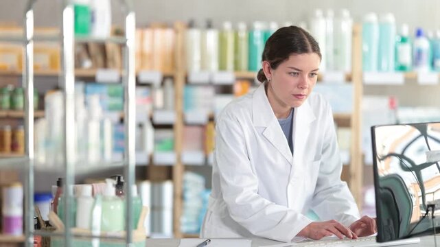 Confident skilled young female drugstore staff member in white coat inputting purchase orders into computer system, managing incoming supplies and ensuring stock availability