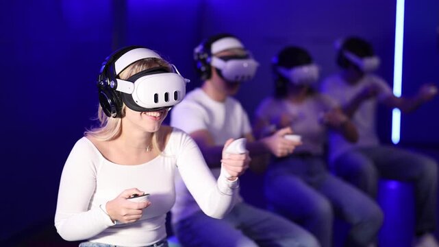 In darkened hall of gaming club, girl in VR helmet takes part in group game. Player overcomes obstacles and reaches goal in parallel reality. Imaginary fantastic world 