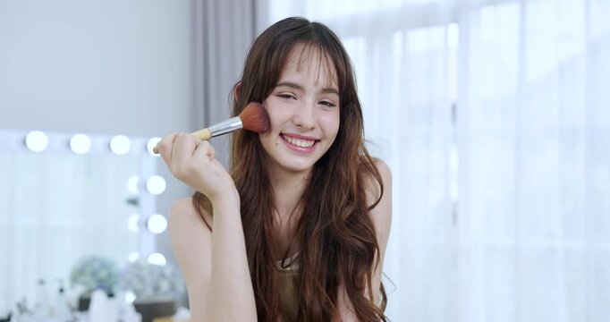 asian female teenager applying blush using makeup brush smiling with closed eyes portraying joy self care beauty moment cozy home skincare routine expressing fresh healthy lifestyle