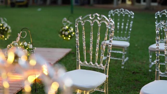Slow pan across transparent acrylic chairs on a grass lawn beside a decorated wedding aisle, with soft bokeh lights and floral accents, revealing elegant seating layout and refined outdoor ambiance