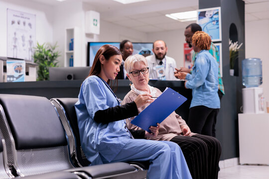 Asian gerontology physician in charge of gerontology area requesting personal data from elderly patient. Medical clinic receptionist personnel attending african american sick young couple of people.