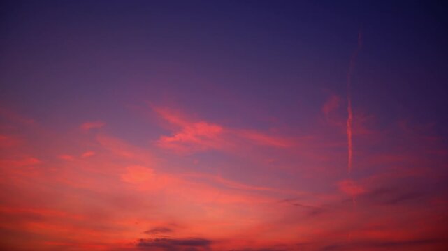 A sunset sky timelapse video footage showcasing wispy cirrus clouds and a thin contrail streak glowing in vivid orange and pink hues, fading into deep blue twilight across a serene evening horizon.
