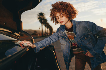 Fototapeta premium Young woman with curly hair wearing rainbow sweater and denim jacket, smiling while opening car trunk outdoors, casual lifestyle, warm sunlight, relaxed mood, natural background
