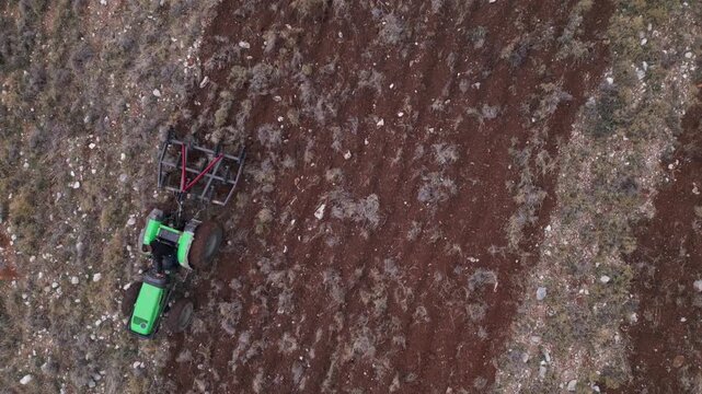 A green tractor with attached tillage equipment churns red soil and dry brush in a top-down view.