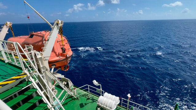 A bright orange fast rescue boat secured on its launching davit on the deck of a commercial vessel, with a background of choppy ocean waves during transit in windy maritime conditions