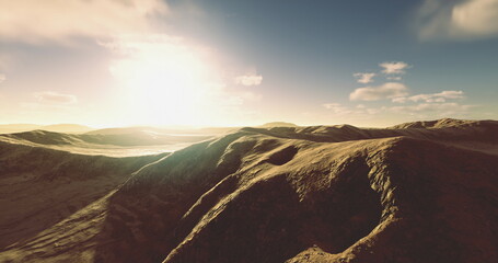 Golden sunlight bathes the gently rolling hills, casting long shadows across the rugged terrain as clouds drift lazily in the bright blue sky. A serene moment in nature captured at dawn. © icetray