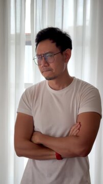 Unhappy and Worried Asian Thai Man wear glasses, standing near window while thinking and considering,  minimal white cozy house.
