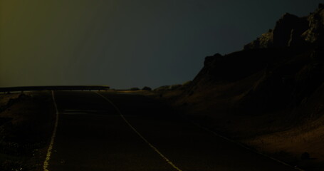 A winding road stretches into the darkness, flanked by rugged rock formations. Dim lighting casts an eerie glow over the landscape, creating a mysterious atmosphere at night. © icetray