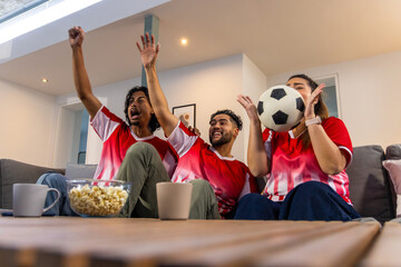 Naklejka premium Friends wearing red white jerseys cheering, watching match on sofa with soccer ball, popcorn, mugs