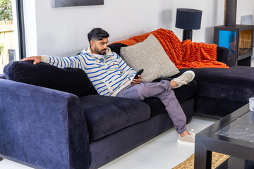 Fototapeta premium Indian man in his 30s reclining on dark navy sectional sofa at home, holding smartphone