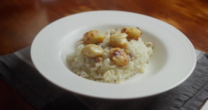 Serving creamy seafood risotto with seared scallops onto white plate. Close up of hot homemade rice dish being plated on wooden table, gourmet dinner preparation in natural light.