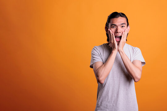 Shocked young man with hands on cheecks and open mouth, news hearing, omg sign. Excited asian teenager screaming, amazed person portrait, medium shot on orange background