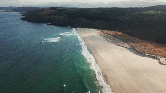 Drone pulling back diagonally over Praia do Rostro in Galicia, Spain revealing wide sandy beach, Atlantic waves and cloud shadows.