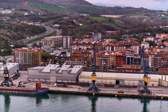 Pasai Donibane, Spain - 10 March 2026: Aerial view of a harbor where the water reflects the cloudy sky, contrasting with the vibrant buildings and industrial cranes.